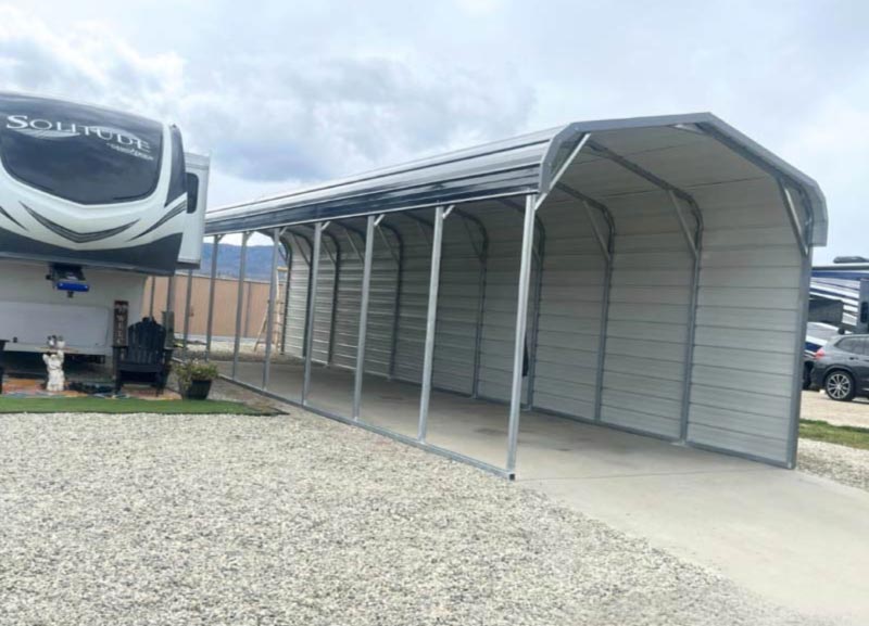 High-clearance regular roof steel RV carport structure with open side panels next to a parked luxury fifth-wheel trailer on a gravel lot.