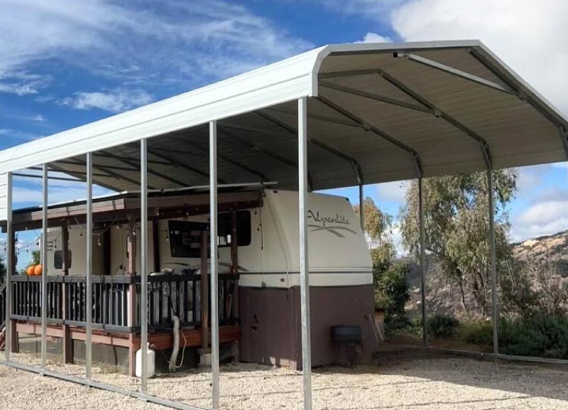 Wide regular-style curved roof metal RV carport installed over an outdoor living patio deck and a parked white travel trailer.