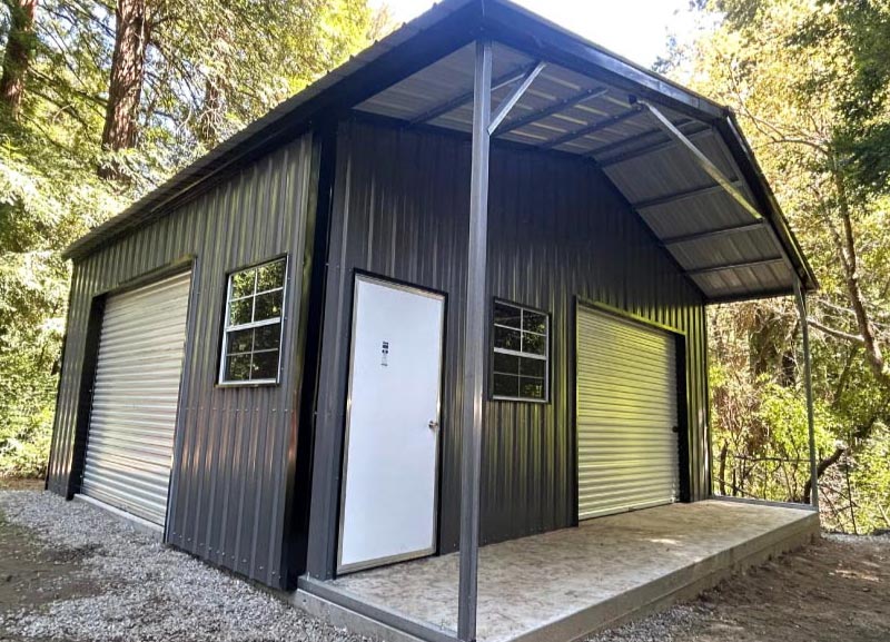 A modern charcoal gray steel utility workshop nestled in a wooded setting, featuring a covered front porch canopy, secure roll-up garage doors, a white walk-in entrance door, and framed windows.