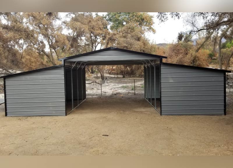 Symmetric gray metal barn structure with a central open-drive carport and dual matching lean-to side storage units on a dirt ground surface.