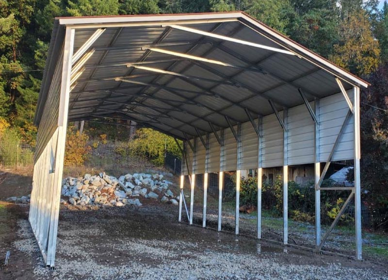 Tall open-frame metal RV shelter featuring an industrial A-frame roof configuration and integrated concrete anchor footings in a wooded area.